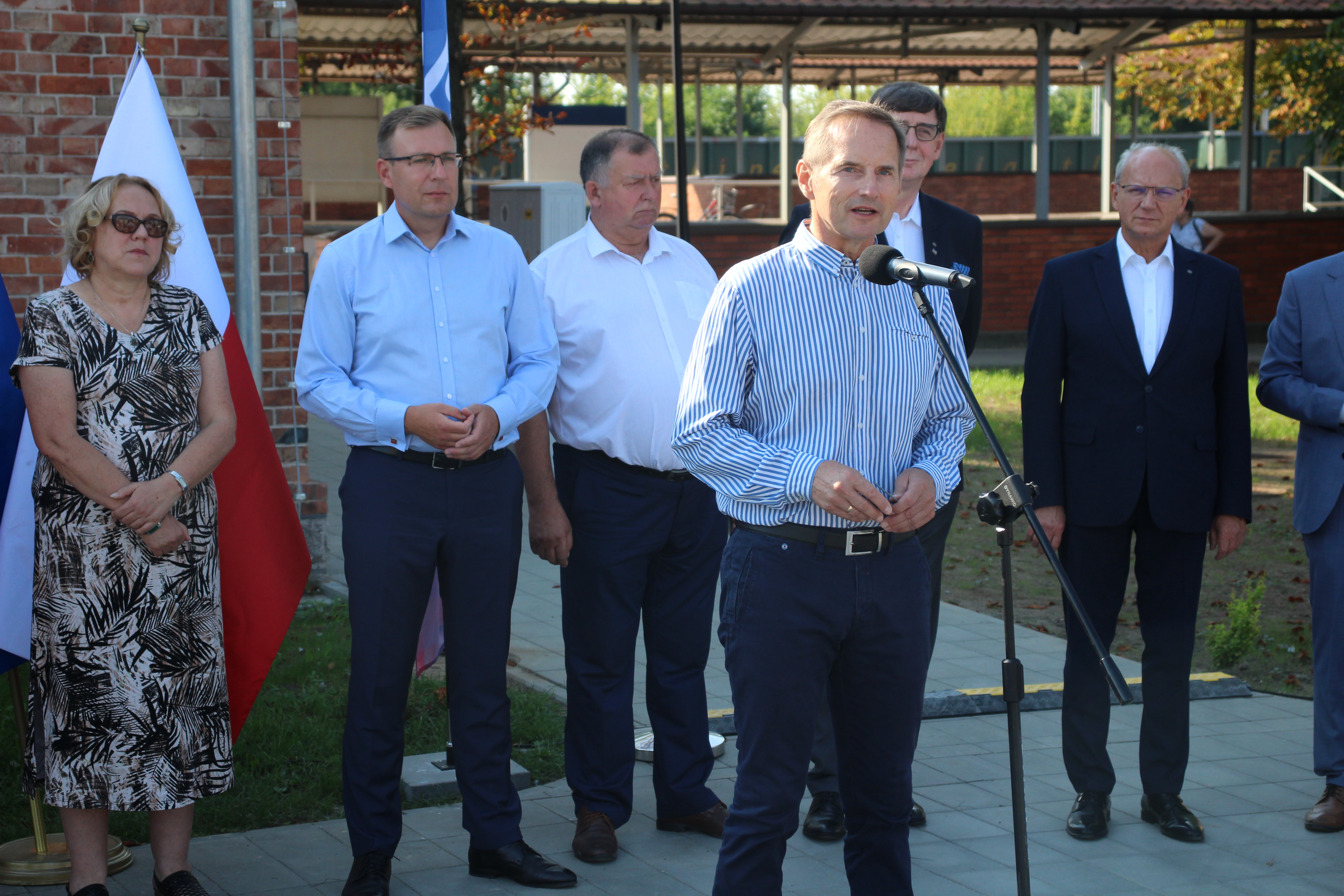 Burmistrz Piotr Osiecki przemawia na&nbsp;tle wyremontowanego dworca. Za nim kilka os&oacute;b