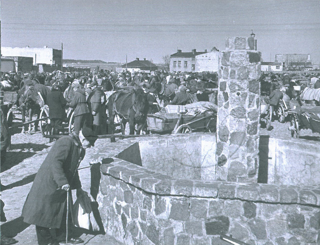 Zdjęcie przedstawia starszego mężczyznę przy kamiennej fontannie. W tle odbywa się targ - widać konie, wozy i&nbsp;wielu ludzi